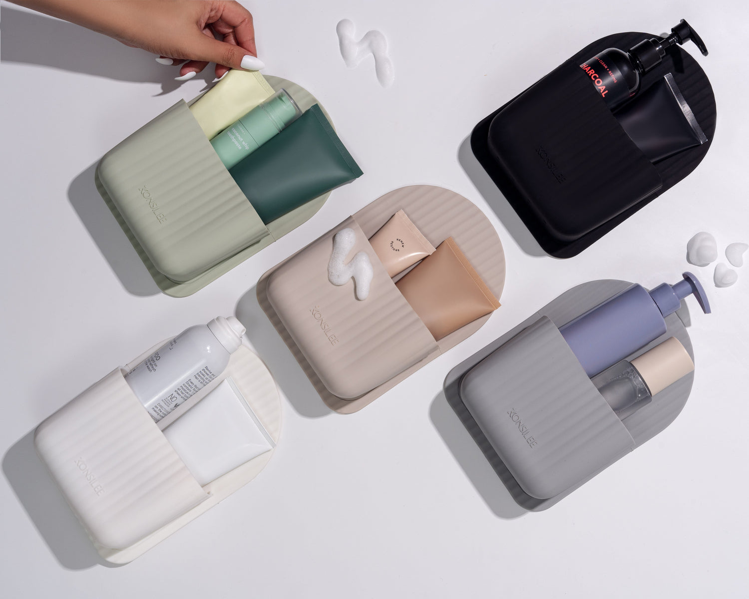 A top-down view of five arch-shaped silicone bathroom organisers in neutral colours: sage green, light beige, black, white, and light grey. Each organiser features a fluted detail and embossed Konsilee logo. The pouches are holding various skincare products like tubes and bottles, arranged artistically on a white surface with a hand reaching for one of the bottles.